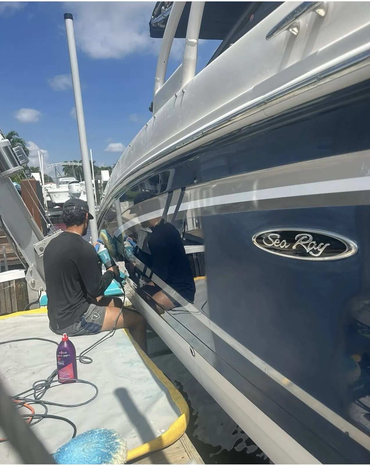 Detailer compounding the blue hull of a Sea Ray at a dock in Boca Harbor, Boca Raton, with a bottle of heavy compound beside him