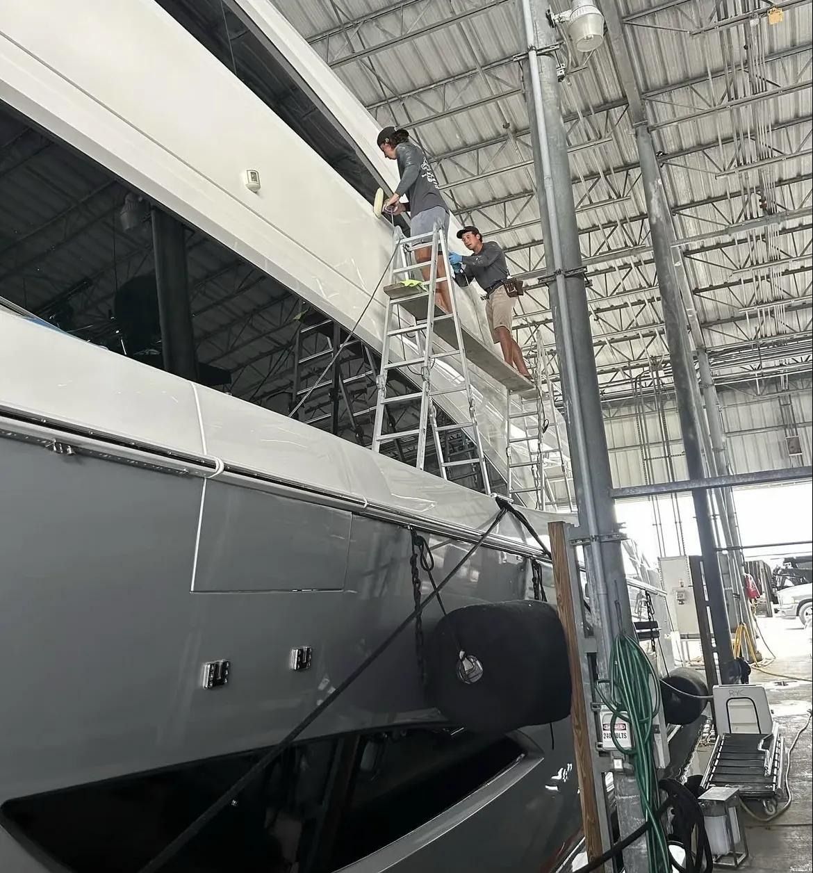Two detailers on a scaffold polishing a large yacht inside a covered shed at Bradford Marine in Fort Lauderdale